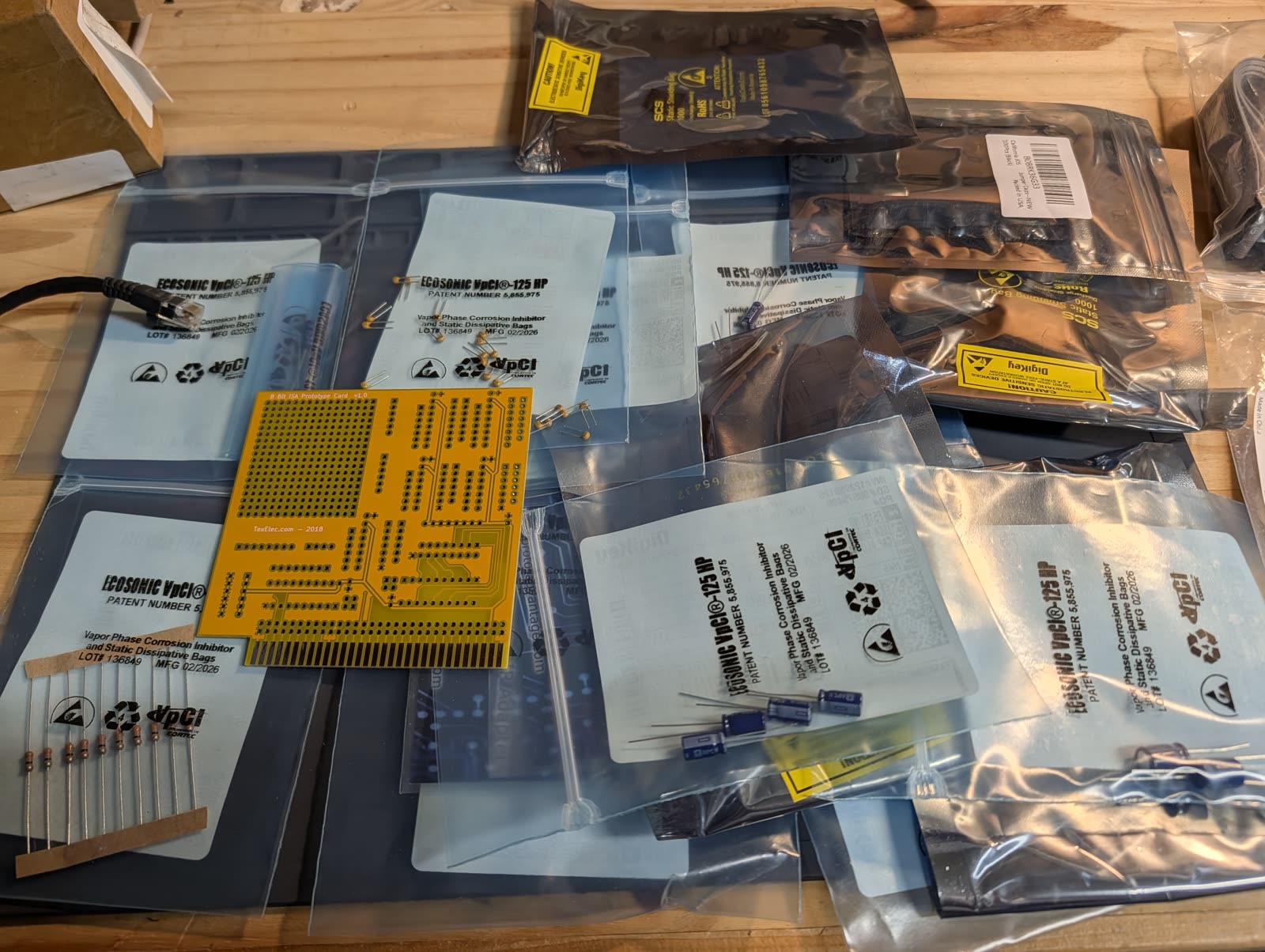 Photo: NetISA parts arrival on a workbench. A TexElec 8 Bit ISA Prototype Card v1.0 (yellow PCB with ISA edge connector) surrounded by EcoSonic VpCI-125 HP anti-static bags containing electrolytic capacitors, ceramic capacitors, resistors, and diodes, along with DigiKey SCS static shielding bags and a ribbon cable.